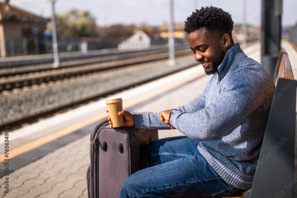 Obraz premium Happy man with suitcase and coffee looking a clock while sitting on a bench at the railway station.