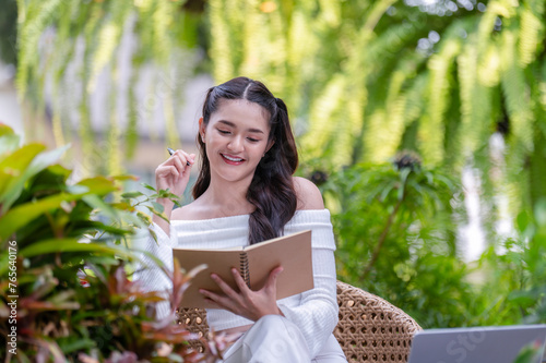 Young woman working remotely using a laptop and taking notes in a notebook Think and come up with project ideas in a relaxed atmosphere at an outdoor cafe.