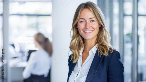 Confident Blonde Businesswoman in Blue Suit, Leading Her Team with a Radiant Smile in Office Setting, Businesswomen Working in Background, Professional Leadership Concept. 