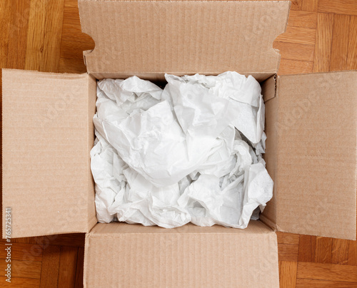 Cardboard box filled with shipping packaging paper on a wood floor, seen directly from above in flat lay style. Freight transportation and e-commerce concept