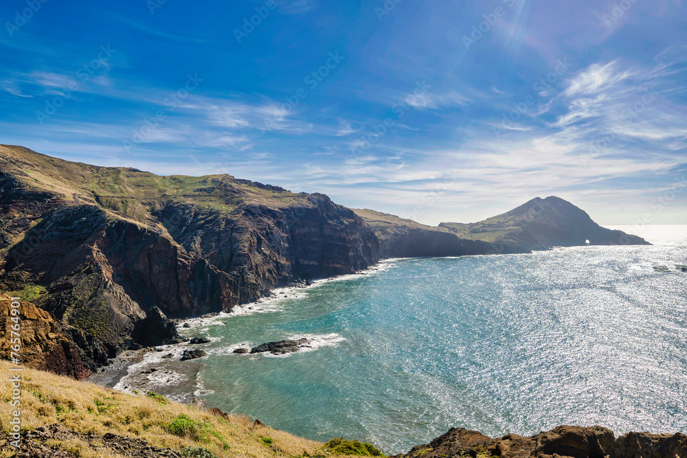 The photo depicts a cliff overlooking a bay and mountains in Madeira. The sky is blue, and the water is blue. In the foreground, there are trees and bushes. A person stands on the cliff.