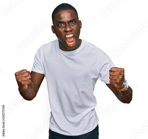 Young african american man wearing casual white t shirt angry and mad raising fists frustrated and furious while shouting with anger. rage and aggressive concept.