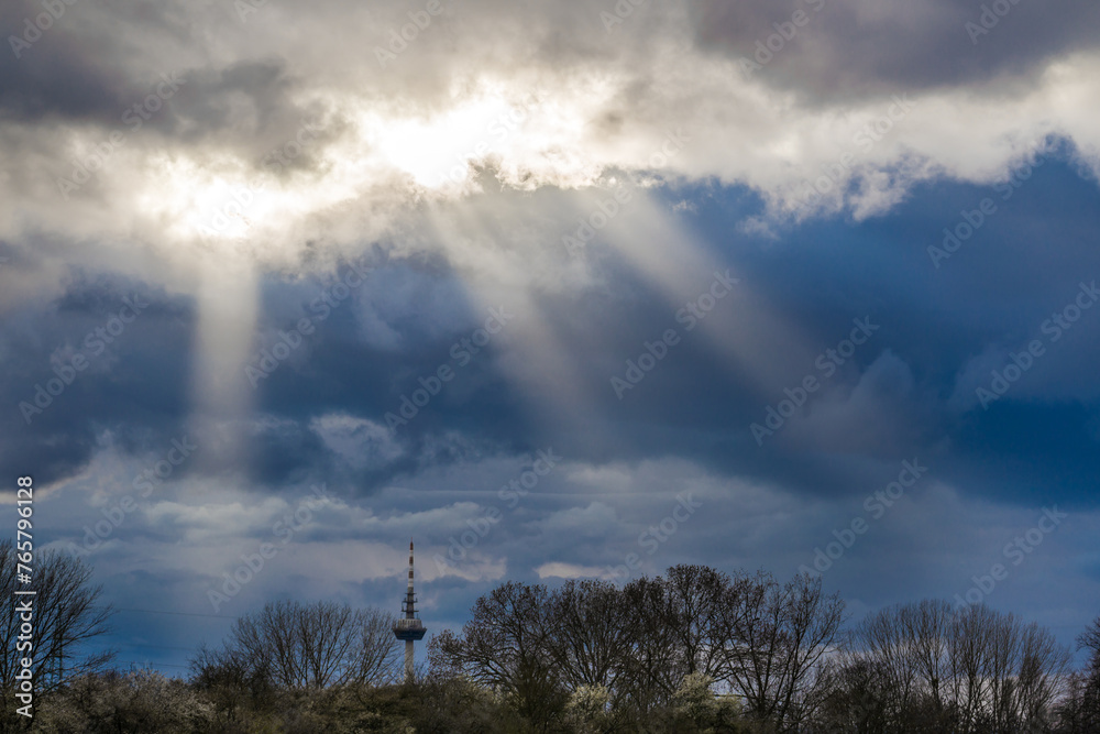 Sun rays in the dark blue sky over the trees and the TV tower, dramatic sky