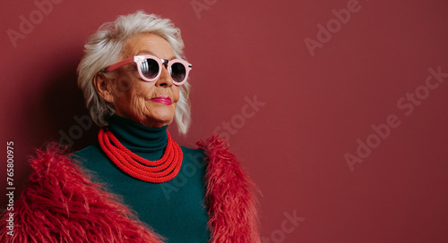 Fashionable elderly woman in stylish eyeglasses looking cool and trendy while standing on red background