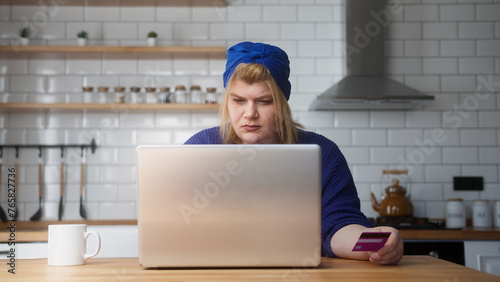 Overweight woman enter credit card number on laptop for shopping online, makes secure easy distant electronic payment while sitting in kitchen at home 