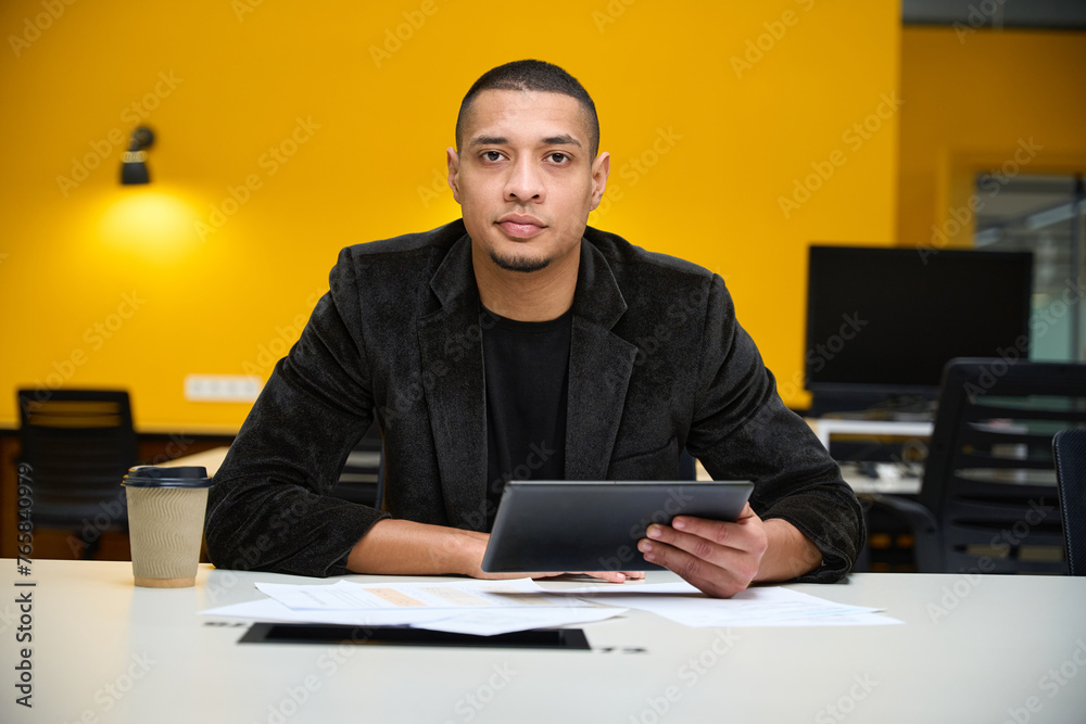 Fototapeta premium Serious young male employee sitting at table and holding tablet