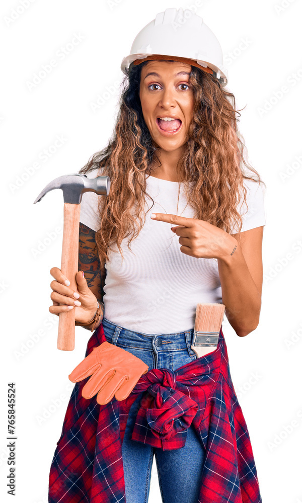 Young hispanic woman with tattoo wearing hardhat and builder clothes holding hammer smiling happy pointing with hand and finger Young hispanic woman with tattoo wearing hardhat and builder clothes holding hammer smiling happy pointing with hand and finger