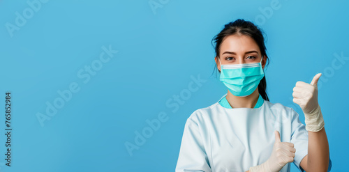 Young pretty female nurse, assistant in a medical coat, mask and gloves show thumb-up fine gesture on blue background. Health care recommendation, medical service, insurance concept