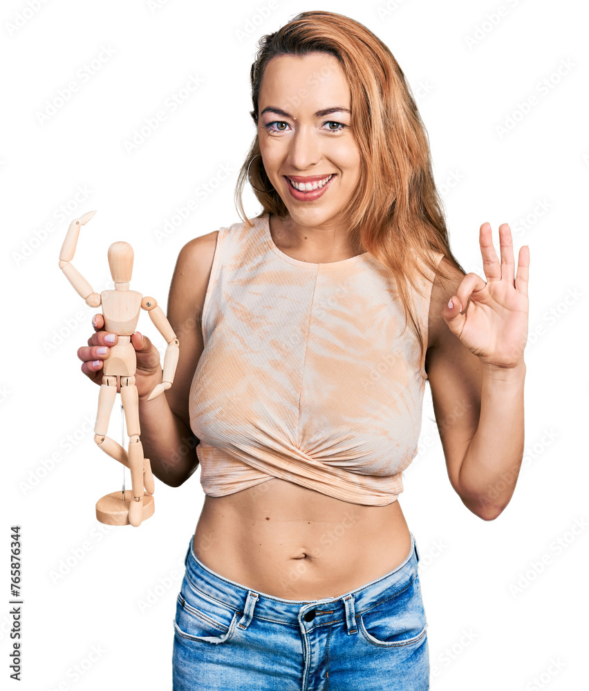 Young caucasian woman holding small wooden manikin doing ok sign with fingers, smiling friendly gesturing excellent symbol Young caucasian woman holding small wooden manikin doing ok sign with fingers, smiling friendly gesturing excellent symbol