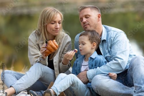 Caucasian mother, father and daughter are on a picnic in a forest on a lake background during fall, eating croissants. Happy family time © Irina Mikhailichenko