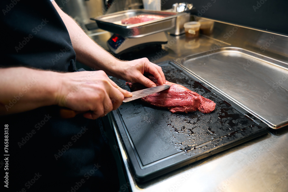 Partial male chef cutting meat fillet with knife on chopping board at table
