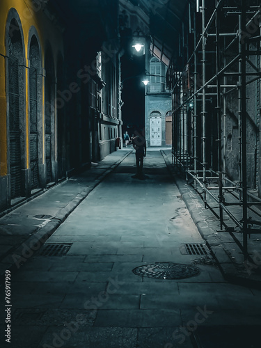 Rua estreita à noite com pouca iluminação e prédios antigos com um homem caminhando.