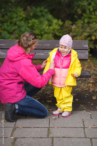 A mother is putting a yellow rainjacket on her little girl and a girl is holding rowanberries in her hand. Pink and yellow kids outfit. Vertical image.