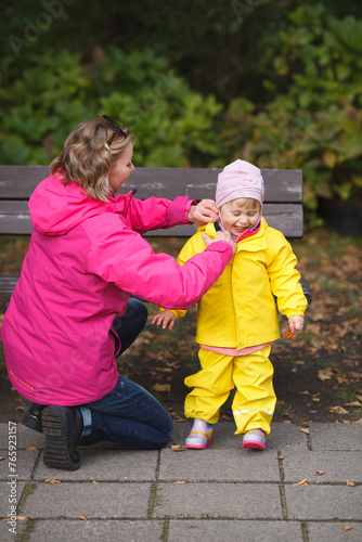A mother is wearing a yellow rainjacket on her little girl and a girl is smiling and holding rowanberries in her hand. Pink and yellow kids outfit. Vertical image.