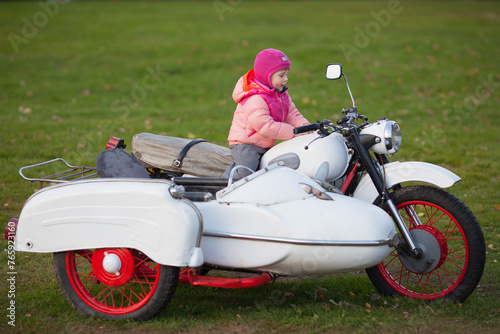 A little girl is sitting on a white vintage sidecar three wheel motorcycle isolated on green grass background. Horizontal image