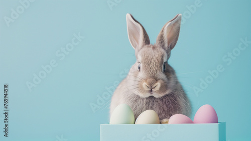 Cheerful Easter Bunny Greeting Card: Adorable Rabbit Sitting in a Gift Box Surrounded by Pastel Easter Eggs, Creating a Playful and Whimsical Holiday Concept Against a Soft Blue Background. © CosmoJulia