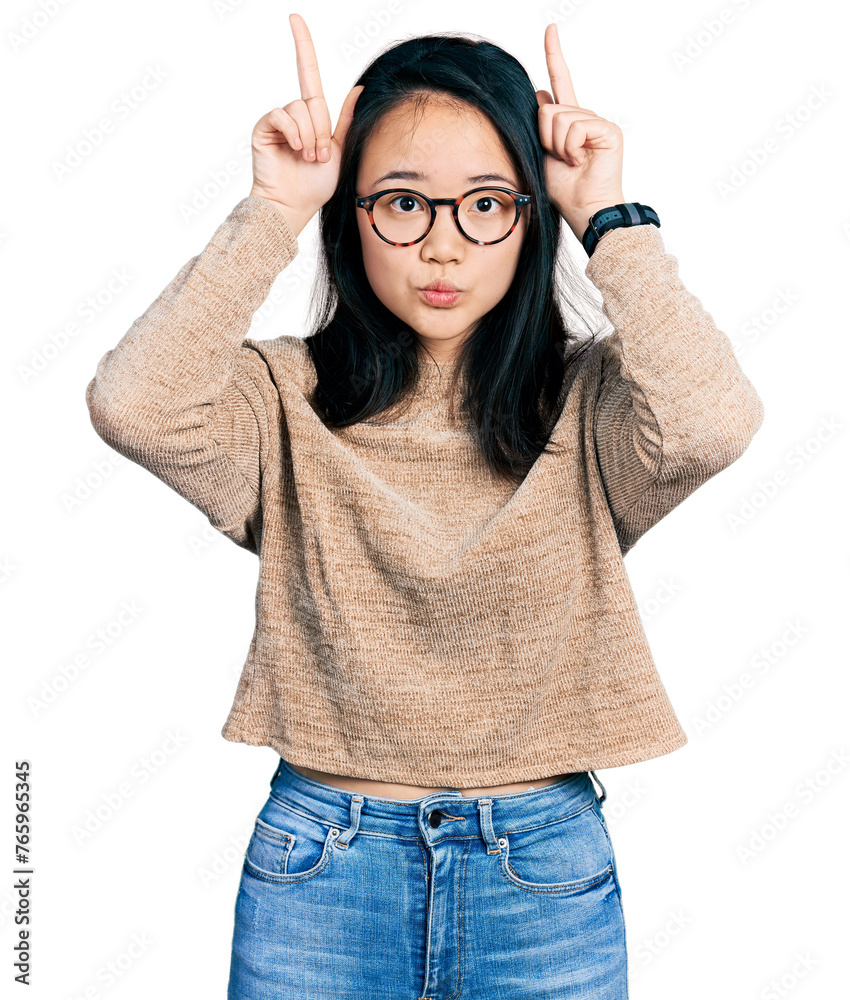 Young chinese woman wearing casual sweater and glasses doing funny gesture with finger over head as bull horns Young chinese woman wearing casual sweater and glasses doing funny gesture with finger over head as bull horns