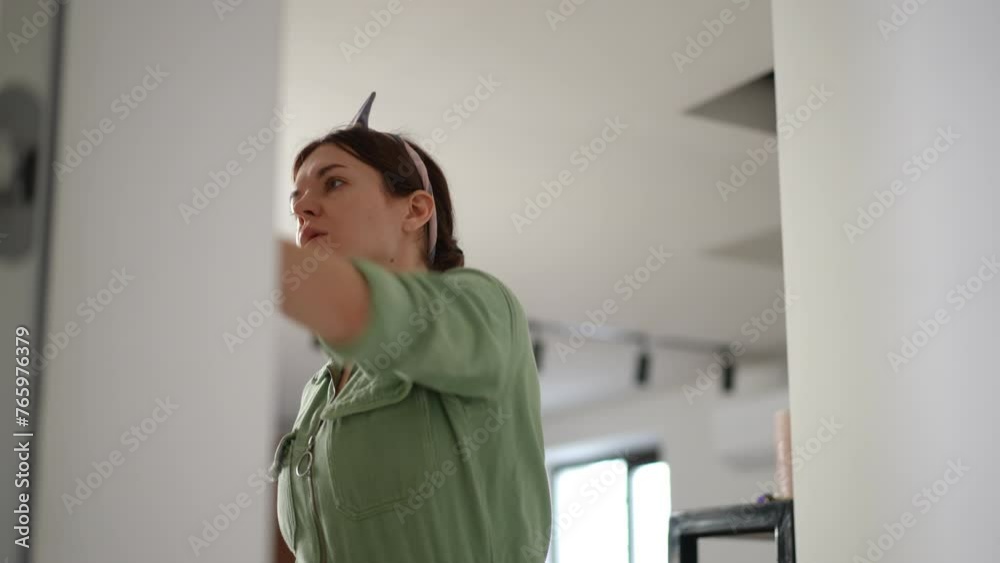 A tired woman in a green overalls has finished painting the wall with a paint roller with white paint, standing on a stepladder in the apartment corridor A tired woman in a green overalls has finished painting the wall with a paint roller with white paint, standing on a stepladder in the apartment corridor