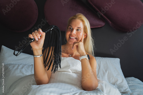 A young blonde girl of European appearance in a bed in the bedroom with a whip in her hand.