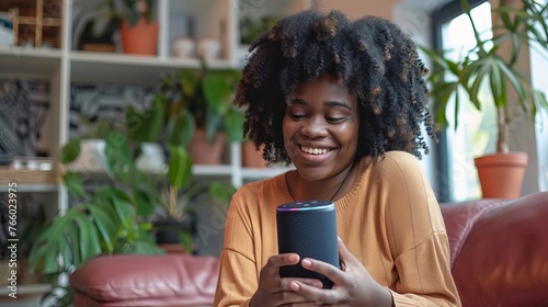 Black woman using her virtual assistant smart speaker to connect her phone. A content black woman at home with a smart speaker. Young, happy woman using voice commands to operate household appliances photo