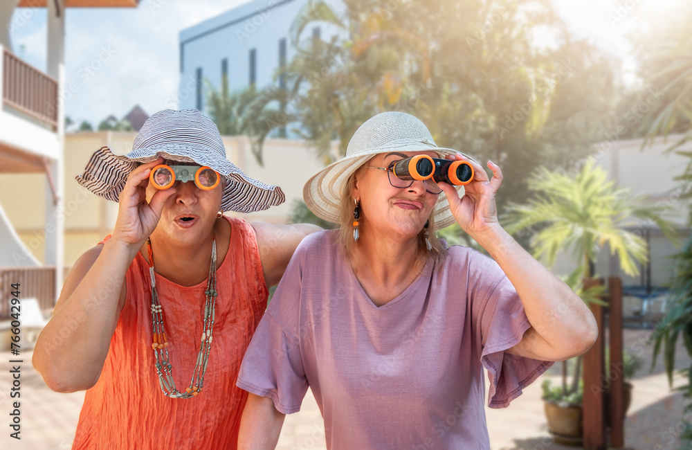 Senior girlfriends in hats and binoculars look in different directions at the resort