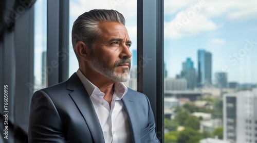 mature optimistic Latino Hispanic businessman executive CEO in corporate modern office thinking contemplating and looking out window skyscraper cityscape daytime with room for text