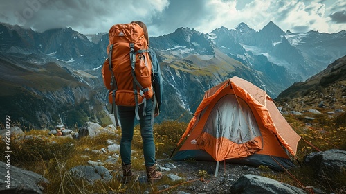 Hiker standing at orange tent in front of camp and carrying backpack on mountain