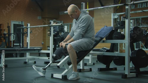 Motivated athlete with prosthetic leg corrects prosthesis and starts to train with empty barbell in modern gym. Fit man with physical disability does exercises using professional sports equipment. photo