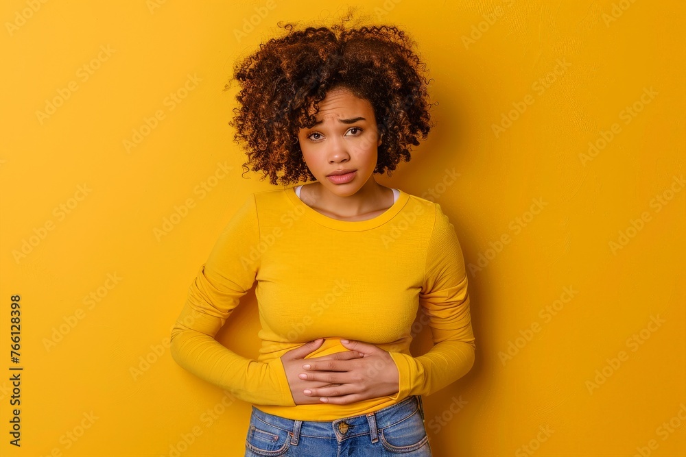 a worried woman in a yellow shirt touching her stomach on yellow studio background - obrazy, fototapety, plakaty 