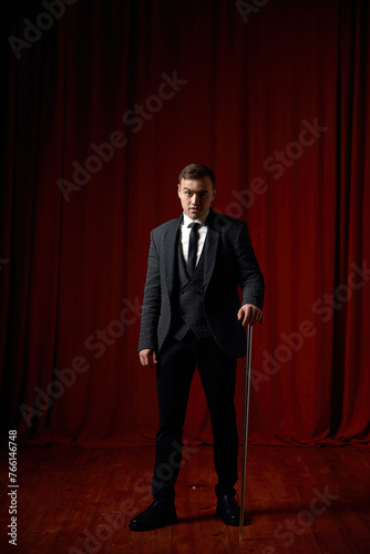 Magician wearing suit standing with cane on stage decorated red velvet curtain