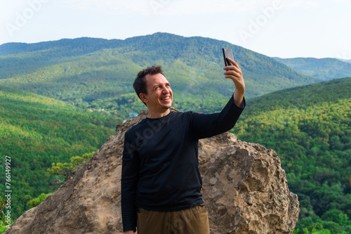 A young smiling male tourist in a black T-shirt takes a selfie on top of a cliff in summer