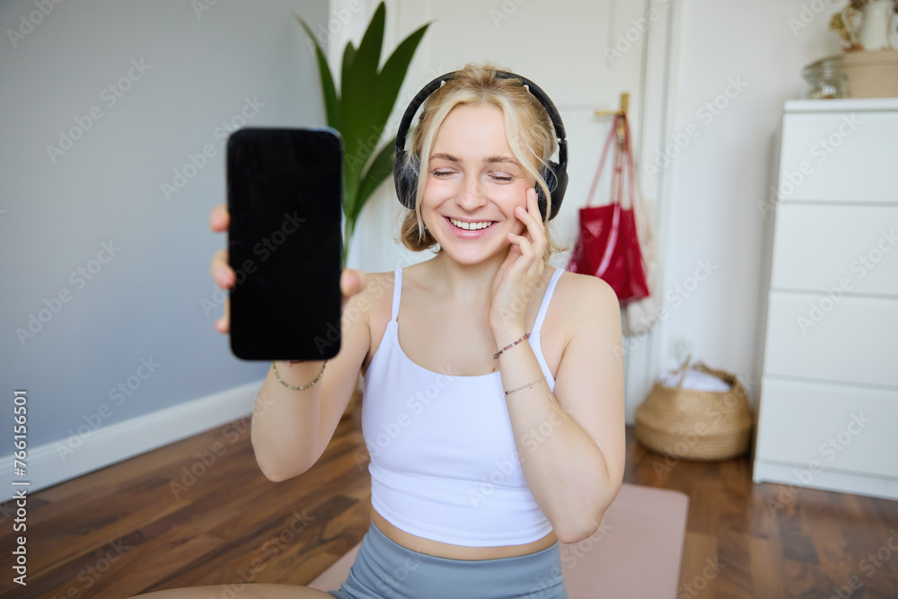 Portrait of smiling, beautiful young woman, showing her smartphone screen, wearing headphones, looking satisfied with sound quality - obrazy, fototapety, plakaty 