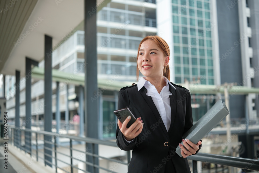 A young Asian businesswomen wearing a suit holding files standing in a big city on a busy downtown street. Young Asian businesswoman using smartphone texting to contact clients. A young Asian businesswomen wearing a suit holding files standing in a big city on a busy downtown street. Young Asian businesswoman using smartphone texting to contact clients.