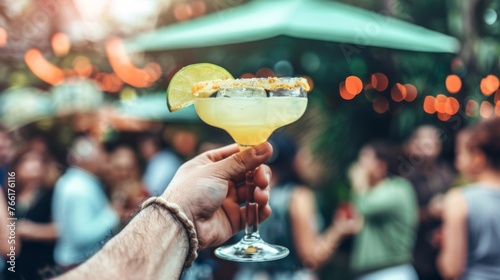 A hand presents a Margarita cocktail, its rim crusted with salt and garnished with a lime, against a blurred backdrop of a social event, evoking the warmth and conviviality of shared moments.