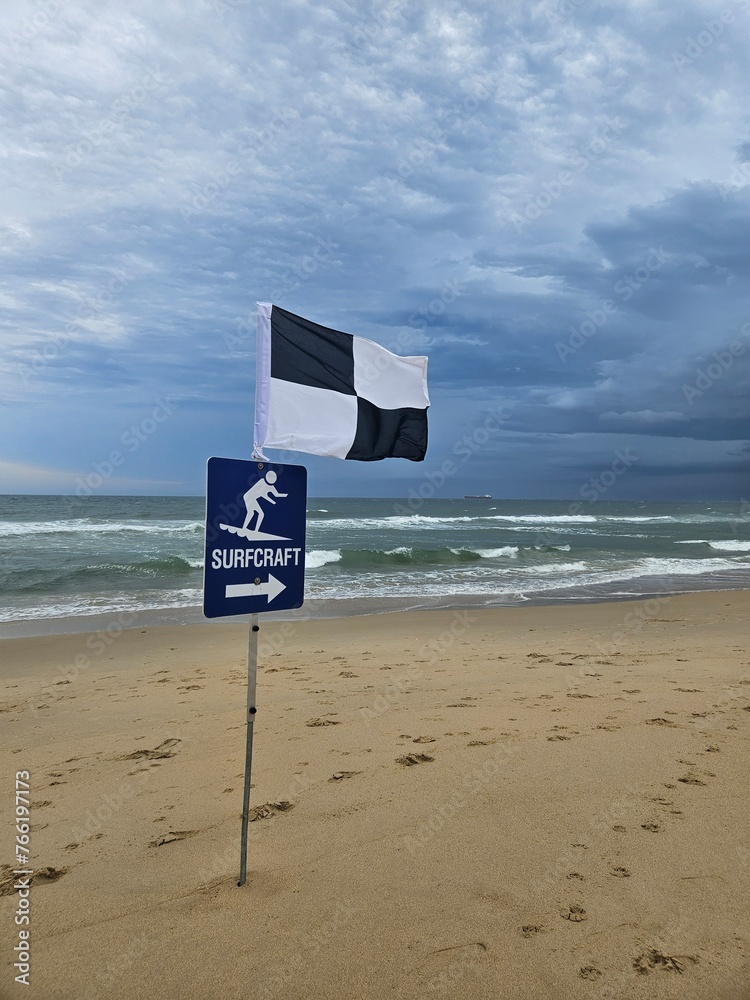 Cloudy Beach Day: black and white flag marks designated surfcraft area, waves gently crashing as footprints scatter across the beach - obrazy, fototapety, plakaty 