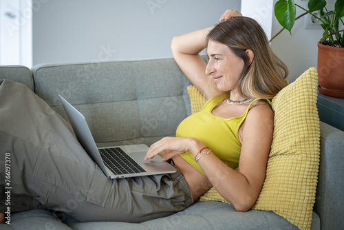 Relaxed freelancer woman online working on laptop, lying on sofa at home. Carefree female in yellow top using computer on couch, resting, watching movie, comfort at living room. 
