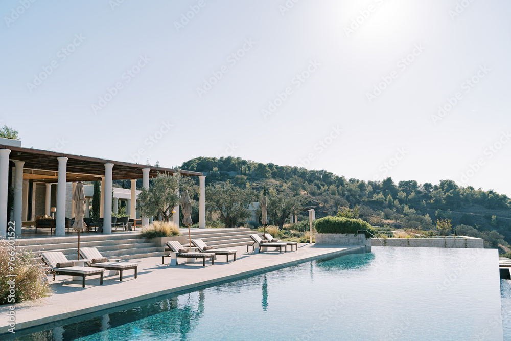 Sun loungers with folded sun umbrellas line the pillared terrace next to the long swimming pool. Hotel Amanzoe. Peloponnese, Greece