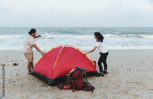 A man and a woman setting up a red and yellow tent on a sandy beach with backpacks nearby  under a cloudy sky  suggesting a camping trip or a leisure activity by the ocean.