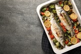 Delicious fish with vegetables and lemon in baking dish on grey textured table, top view. Space for text