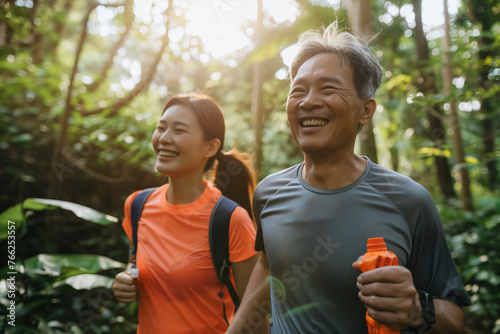 Fototapeta Naklejka Na Ścianę i Meble -  A Asian man and woman are walking through a forest, smiling and holding water bottles. Scene is happy and carefree, as the couple enjoys their time together in nature