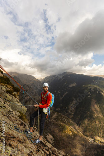 A man in an orange jacket is standing on a rock with a rope attached to him. He is wearing a backpack and he is preparing for a climb. The sky is cloudy
