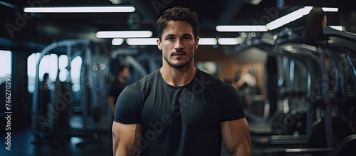A focused man wearing a black shirt is standing inside a gym  surrounded by fitness equipment and weights