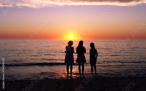 Three friends stand together in peaceful contemplation on a beach, silhouetted against the captivating backdrop of a sunset.