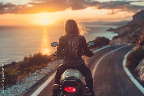 Rear view of a young long haired woman without a helmet riding a motorcycle on an asphalt road near the sea under sunset lights