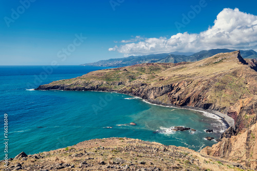 The photo depicts the picturesque landscape of Madeira PR8 Vereda da Ponta de S  o Louren  o  light blue sea  rocky coastline  and green hills under a blue sky.