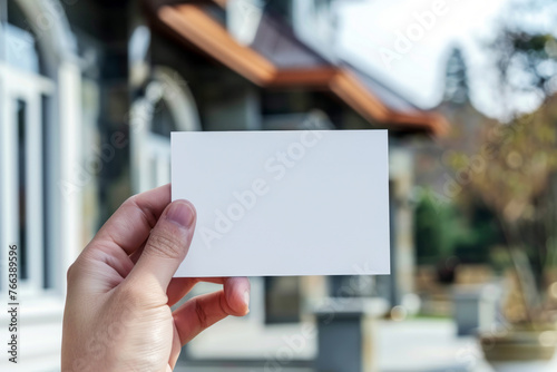 Close-up of Hand Holding Blank White Card, Blurred Residential Building in the Sunny Background © thanakrit