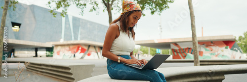 Cute tanned woman with long brown hair in white top and yellow bandana using laptop while sitting on bench. Pretty girl student preparing for classes in outside, Panorama photo