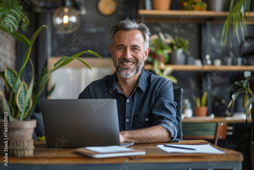 A man is sitting at a desk with a laptop and smiling. Businessman using laptop computer in office space, happy middle aged man boss, entrepreneur, business owner working online on big workplace
