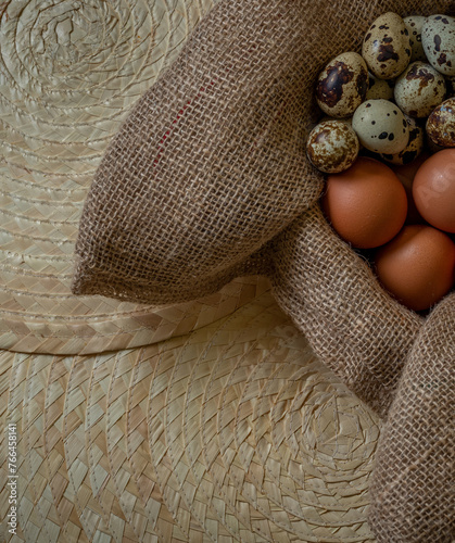 Fresh chicken and quail eggs in a basket on a sack, wooden table.Selective focus