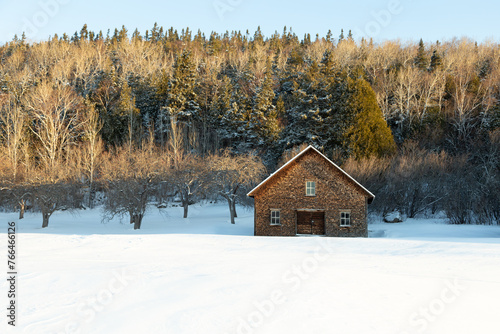 Early morning sunny winter landscape featuring a shingled brown rustic cabin set in fresh snow with mixed trees and mountain in the background, St-André-de-Kamouraska, Quebec, Canada © Anne Richard